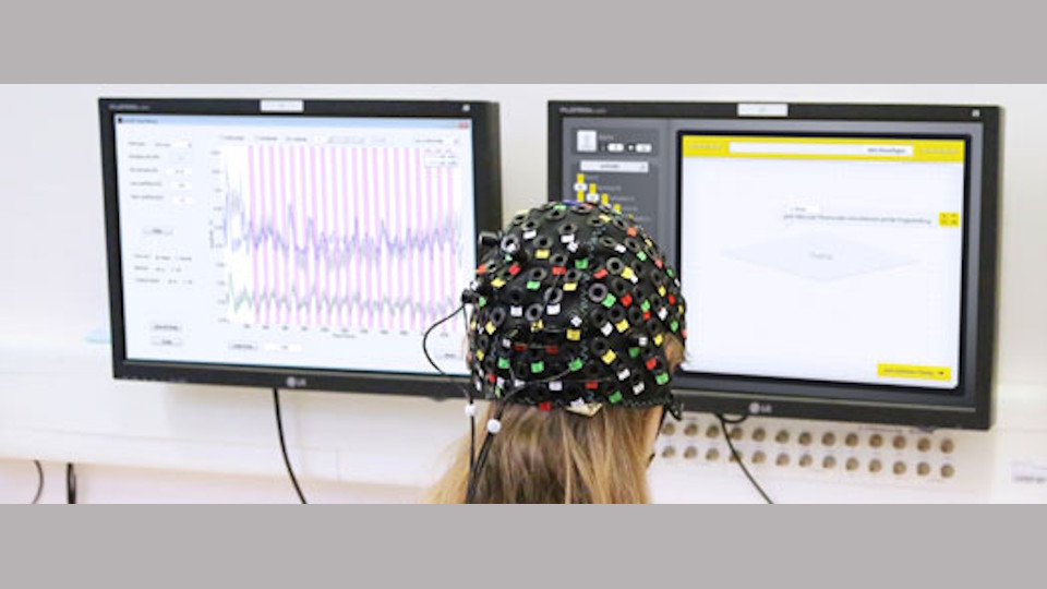 Foto von einer Probandin mit angeschlossener EEG-Kappe schaut auf zwei Monitore